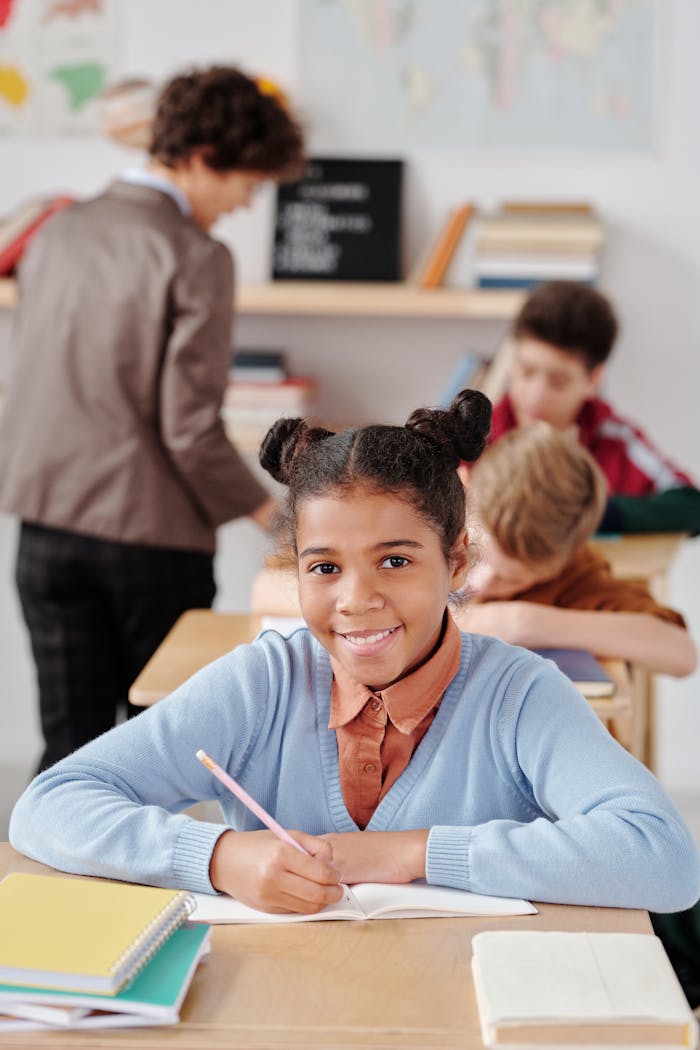 Young girl smiling while writing in a classroom, surrounded by classmates.